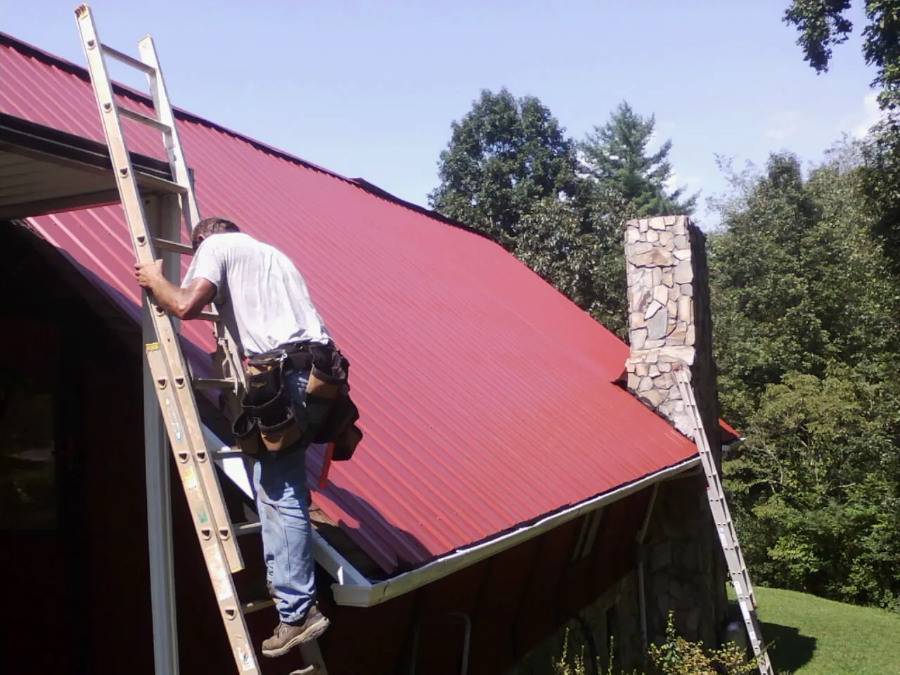 Roofer on a ladder beside a red metal roof and stone chimney — Soffit & Fascia Repair by Roofing Brothers in Glade Valley